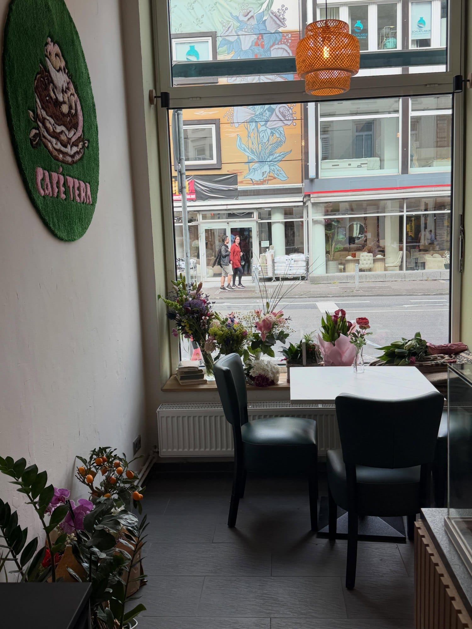 Fensterplatz mit Blumen und zwei Stühlen, an der Wand der runde Logo-Teppich, draußen die Bahnhofstraße