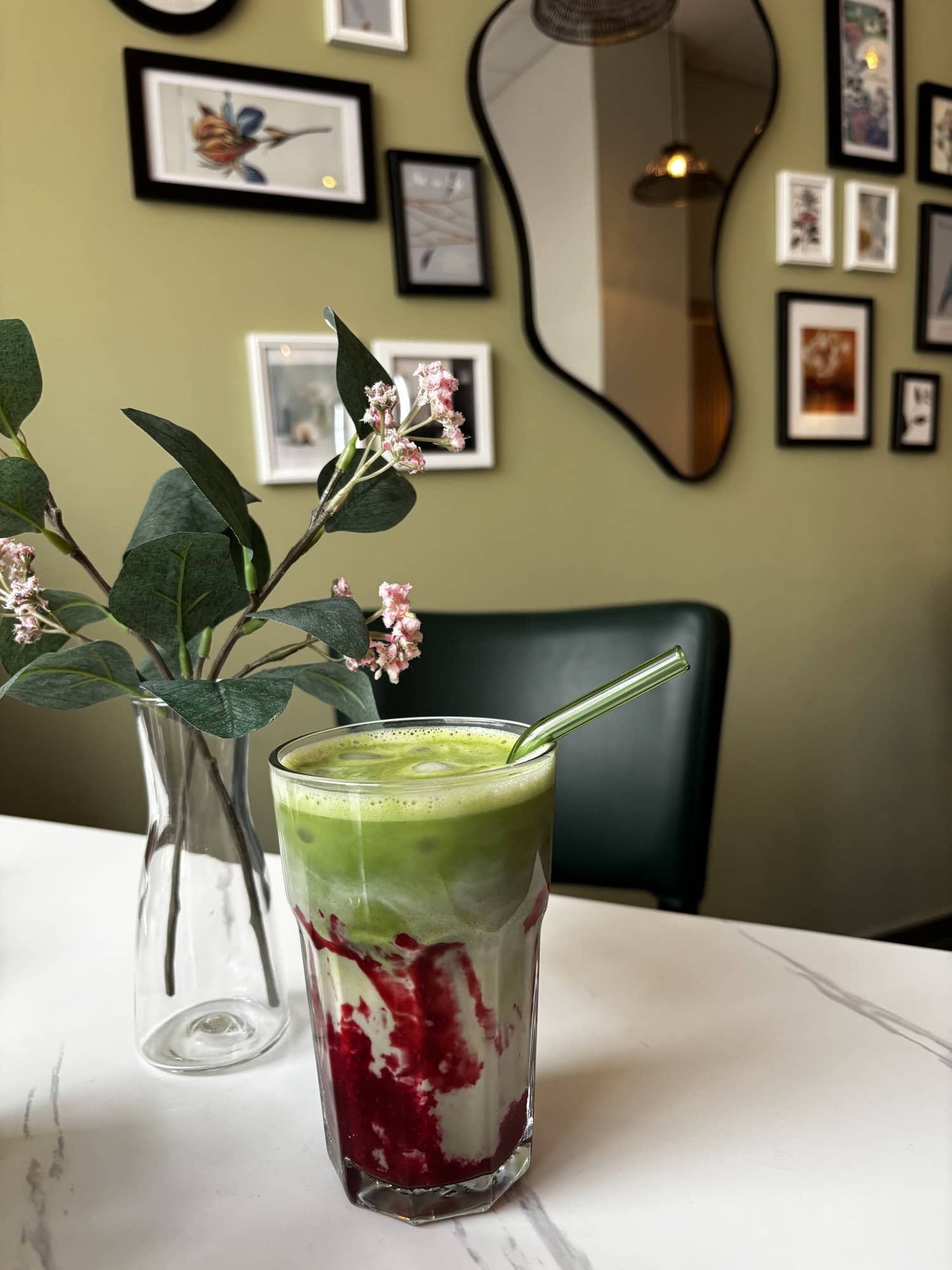 Matcha mit Himbeere im Glas neben einer Vase mit Eukalyptus, dahinter Bilderwand und Spiegel