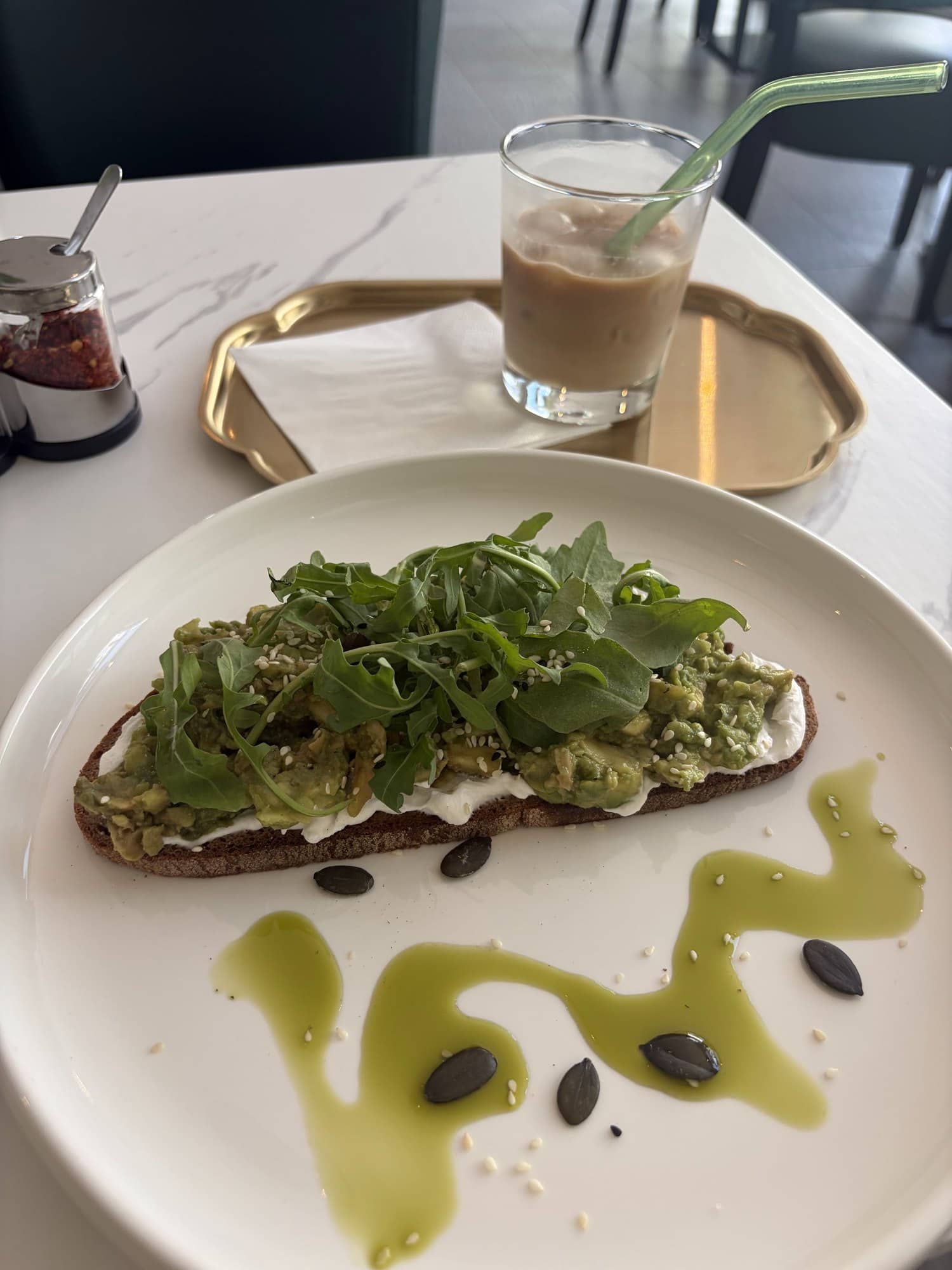 Avocado-Toast mit Rucola und Kürbiskernen, daneben ein Eiskaffee auf einem Messingtablett