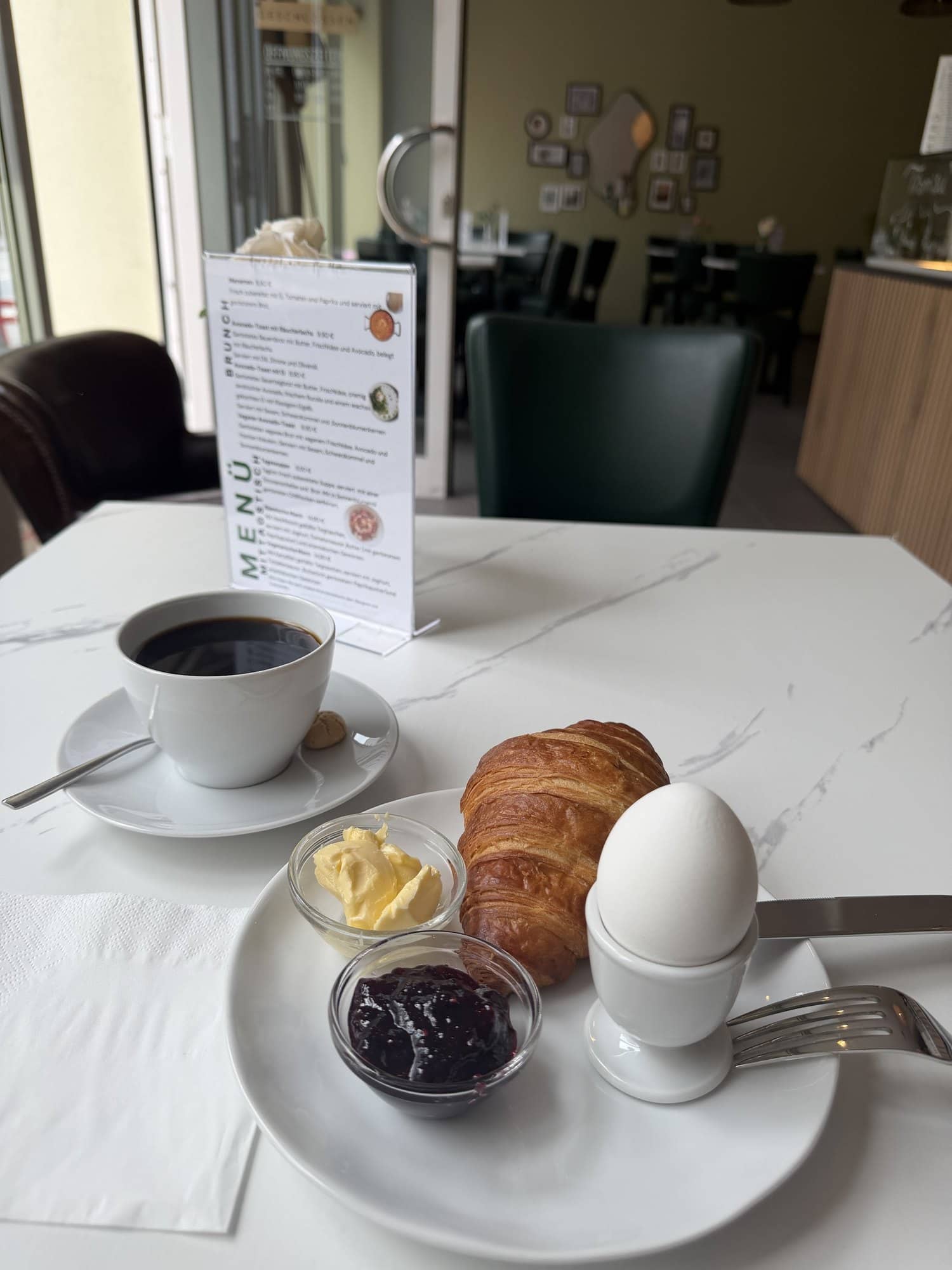 Frühstück mit Kaffee, Croissant, gekochtem Ei, Butter und Marmelade, daneben die Speisekarte