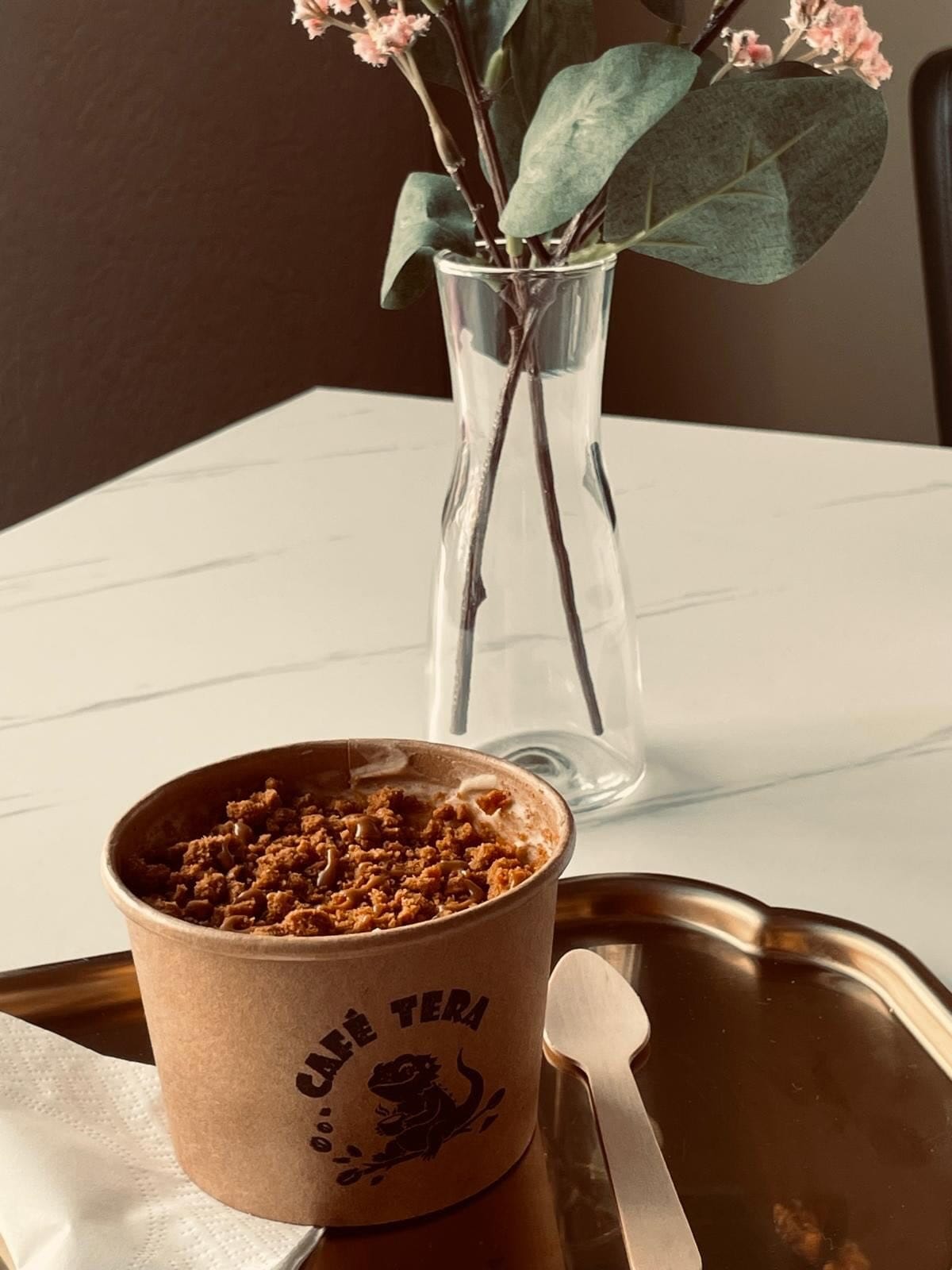 Lotus-Tiramisu im Café-Tera-Becher auf einem Messingtablett, dahinter eine Glasvase mit Eukalyptus und rosa Blüten